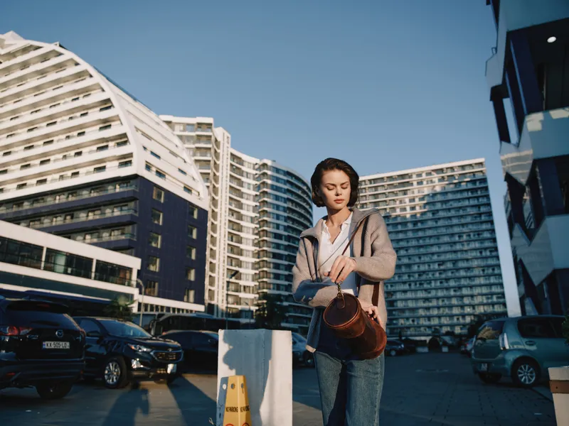 A stylish young woman walks along a city street wearing a casual jacket and bag, enjoying the fresh air as modern high rise buildings rise behind her, creating urban ambiance.