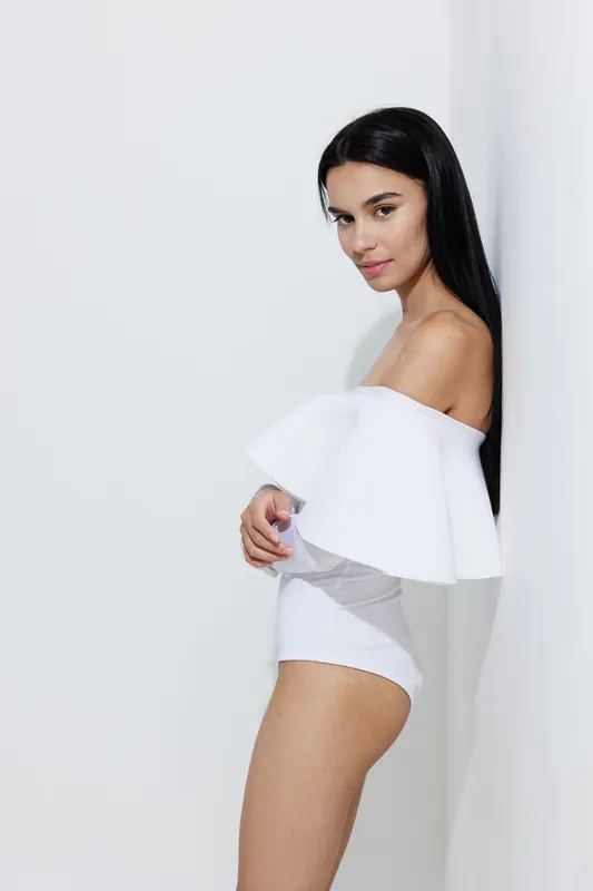 Sport fitness woman with long dark hair wearing a stylish white off-shoulder outfit, posing confidently against a plain light background, embodying health and elegance.