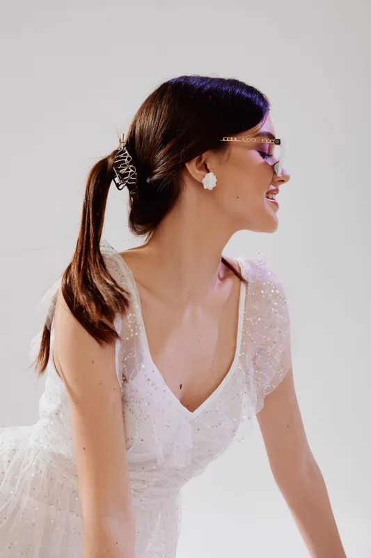 Young woman in a white dress with a ponytail smiles looking sideways. She wears glasses and flower earrings. Studio shot with soft lighting. Concept of woman beauty fashion.
