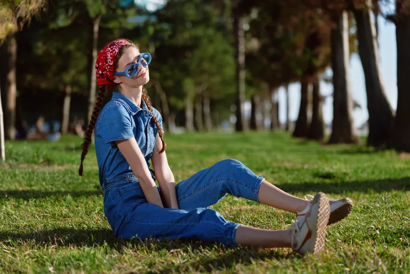 young woman sitting on grass wearing blue denim jumpsuit and red headscarf with sunglasses in sunny park outdoor setting