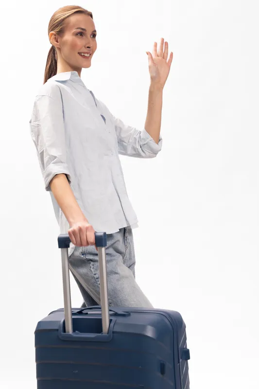 Young woman with suitcase in casual clothes waves hand smiling isolated on white background. Concept for travel, vacation, departure, and tourism with luggage and happy mood.