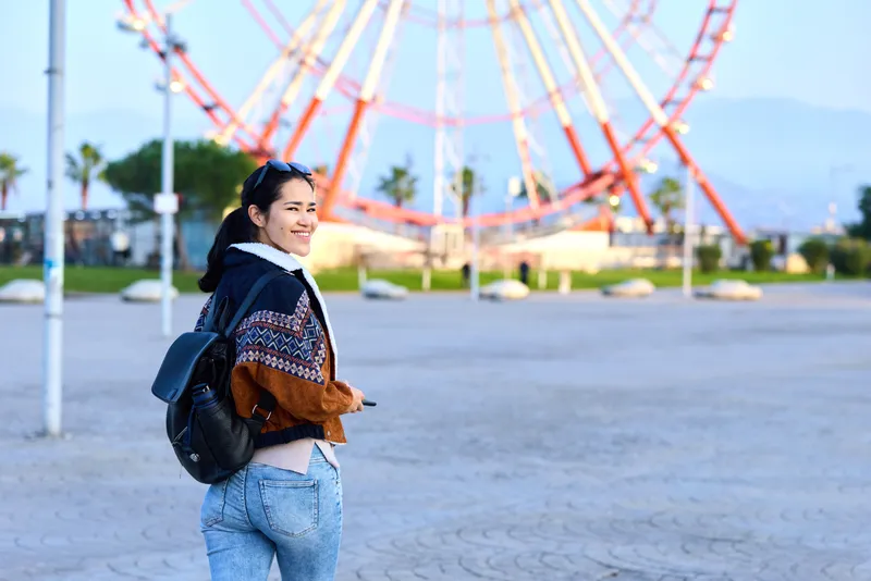 Woman with backpack enjoying a sunny day at an outdoor amusement park near a colorful ferris wheel, casual jacket and jeans, candid moment, cheerful vibe and travel photography