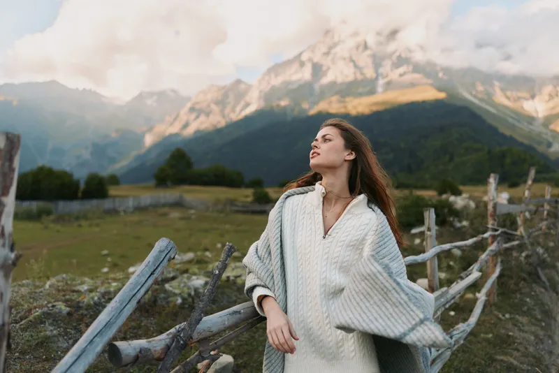 Woman in a cozy poncho stands beside a rustic fence in a mountain landscape, enjoying fresh air and a quiet moment, embracing nature during outdoor lifestyle.