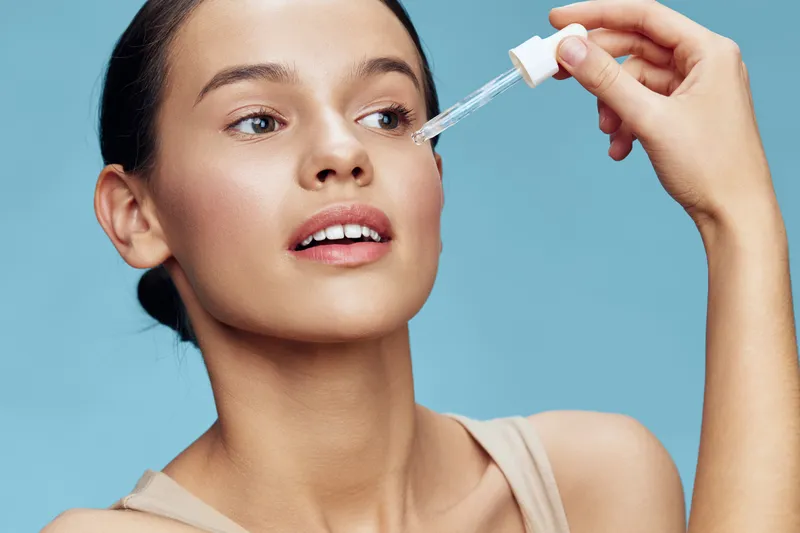 Young woman applying serum to her face, showcasing a healthy skincare routine with a bright blue background and a fresh, confident expression