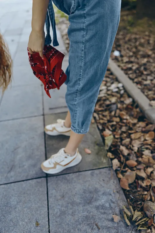 woman holding red scarf in hand wearing casual jeans and sneakers on autumn sidewalk with dry leaves scattered around