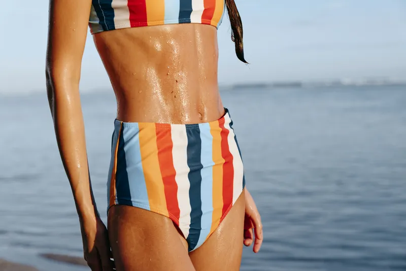Woman in bikini with stripes, closeup outdoors at the ocean in summer, sunlit moment, water droplets on skin, relaxed pose, soft horizon, natural colors, sea breeze, warm air, calm mood