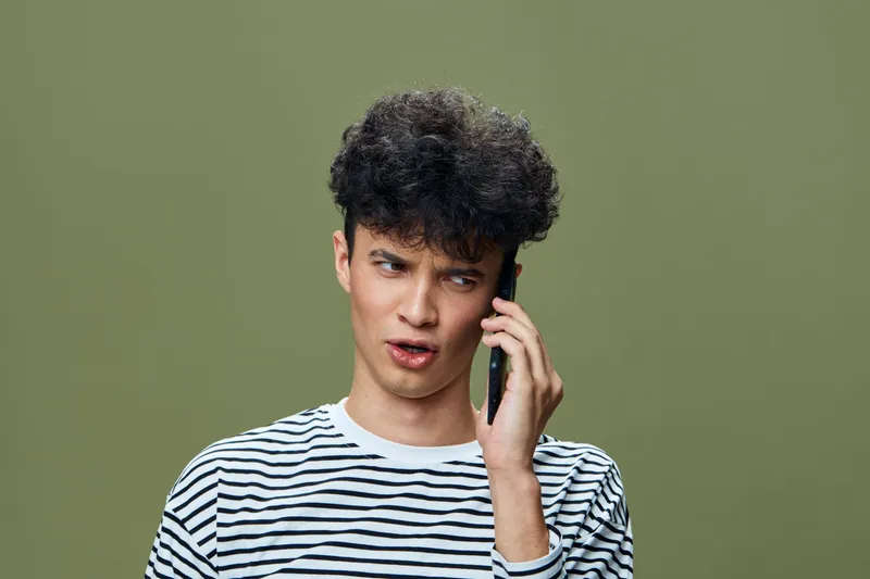 Young man with curly hair wearing striped shirt talking on smartphone with skeptical expression against olive green background, communication and technology concept.