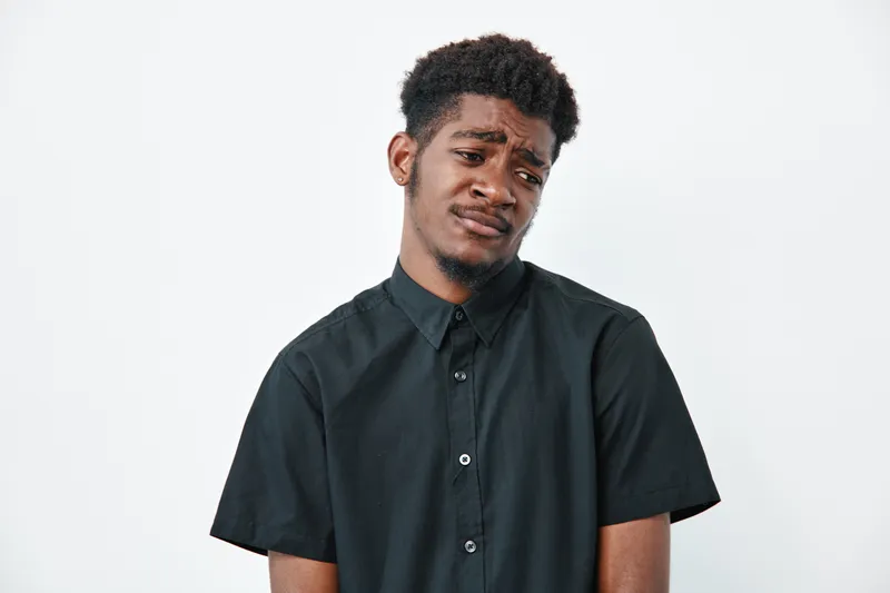 Young black man with dark skin wearing black shirt looking confused or doubtful isolated on white background. Male portrait with afro hairstyle expressing uncertainty or hesitation.