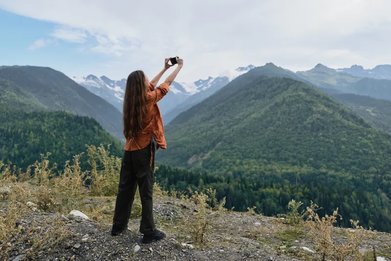 Woman stands on a rocky overlook taking a photograph of the vast mountain landscape with forested valleys and distant peaks under a bright blue sky