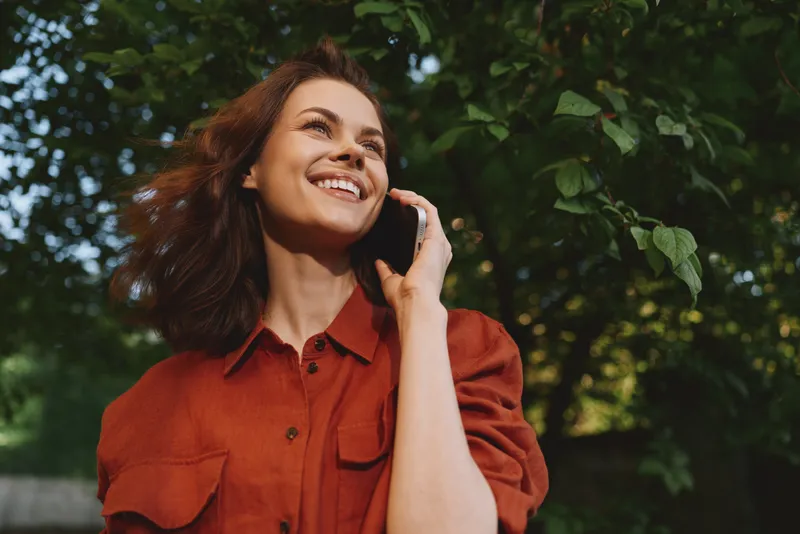 Young woman with shoulder length brown hair talks on a phone in a park, smiling warmly as sunlight filters through green leaves and a light breeze moves her orange shirt