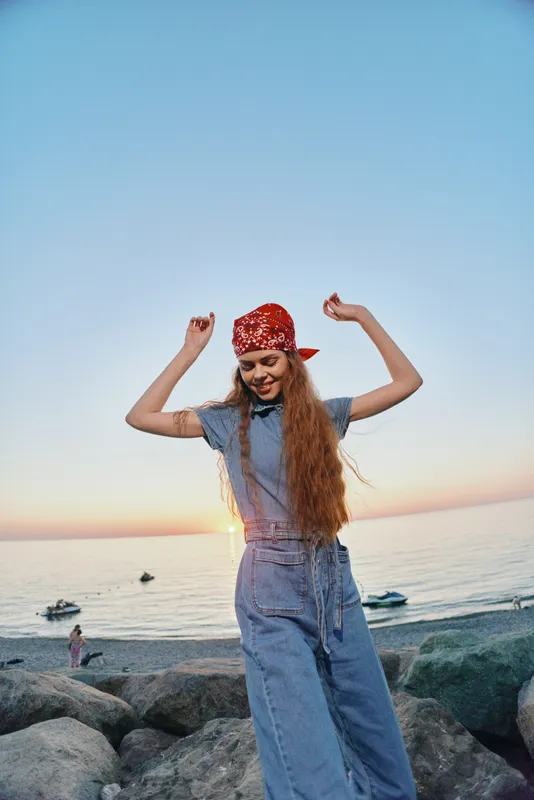 Lifestyle woman in modern boho-western denim street style enjoying sunset by the ocean, wearing a red bandana and relaxed casual outfit in warm film color tones.