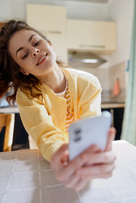 Young woman in a yellow cardigan sits at a kitchen table, taking a casual selfie with a smartphone and a relaxed cheerful expression, at home in natural light