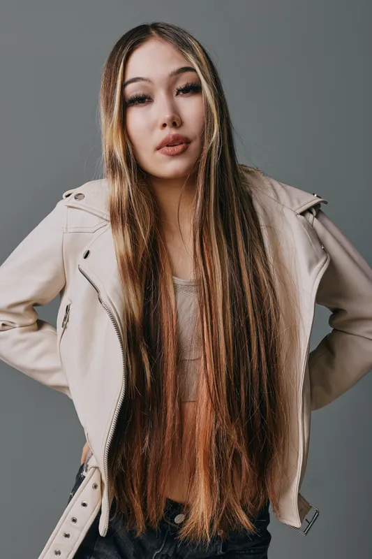 Young woman with long straight hair and casual jacket posing with hands on hips looking confident and relaxed on grey background in studio portrait photo.