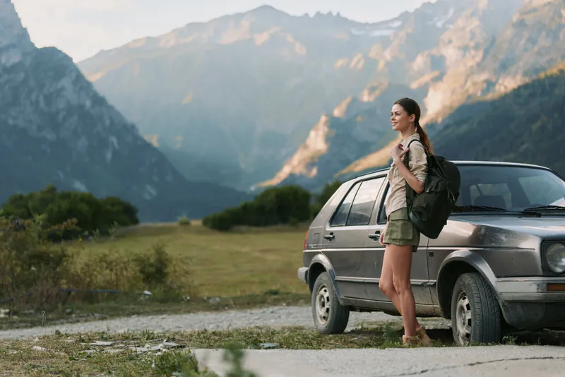 Cheerful woman outdoors, hiking preparation, backpack on her shoulder, sunlit valley backdrop, mountain range in distance, relaxed posture, natural colors