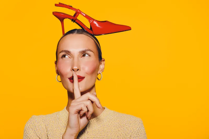 Young woman with red high heel shoes on her head makes shushing gesture on bright orange background. Concept of fashion, silence, and playful mood.
