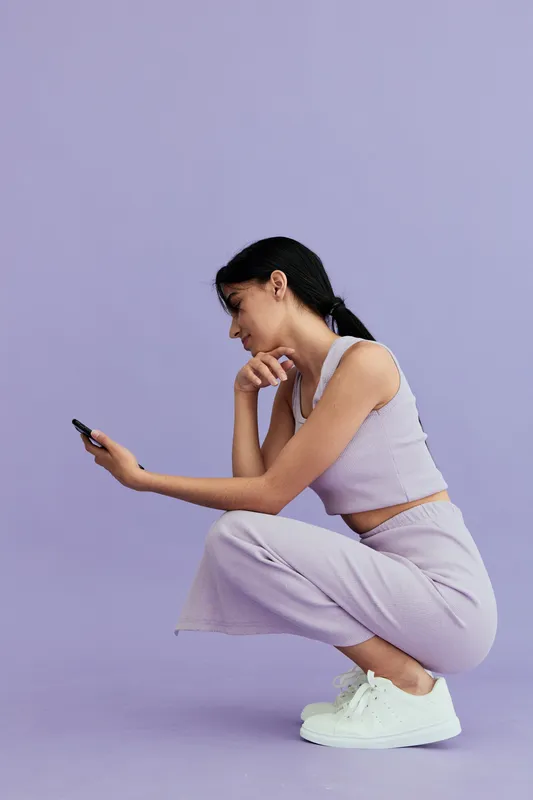 Young woman wearing pastel lilac outfit squatting on violet background, focused on smartphone, casual style, white sneakers, minimalistic color palette