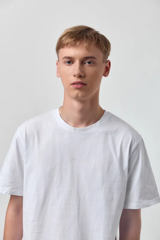 Young man in white tshirt stands against a neutral background, showcasing casual minimalist fashion with a clean look and soft lighting, emphasizing fit, fabric texture, and everyday simplicity
