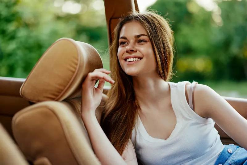 Young woman enjoying a summer day in a convertible car with a joyful smile, wearing casual attire, set against a lush green background.