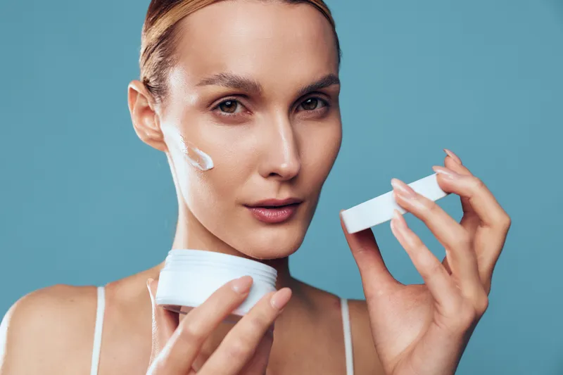 Woman skincare face cream moisturizer beauty spa blue background hygiene, applying facial moisturizer on close portrait, holding cotton pads, calm expression, smooth skin care on clean blue background