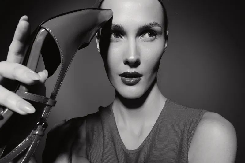 Fashionable white woman with pale skin holds high heel shoe near face in black and white studio portrait.