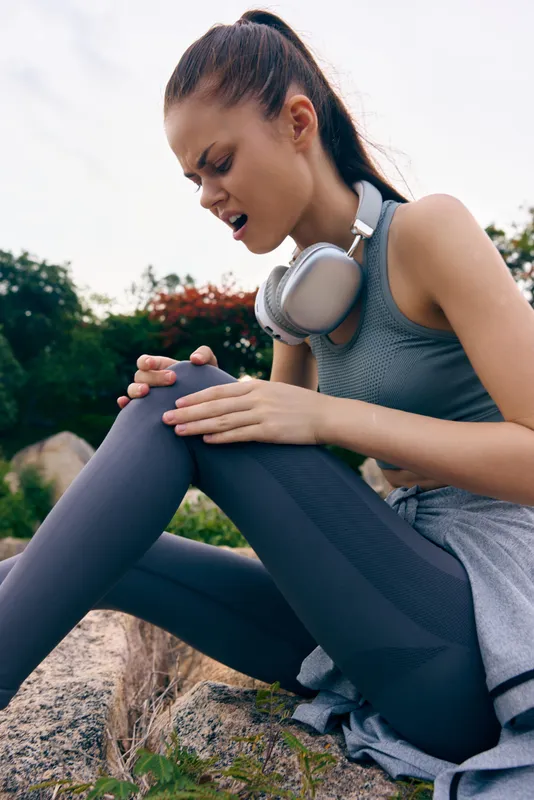 A young athletic woman in workout attire sits outdoors, holding her knee in pain She wears a gray tank top and leggings, with headphones around her neck, reflecting a fitness concept