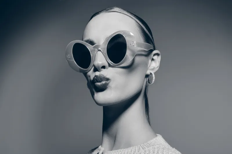 Stylish white woman with pale skin wearing round sunglasses and hoop earrings makes a playful kiss face. Studio portrait in black and white with smooth lighting and modern fashion concept.