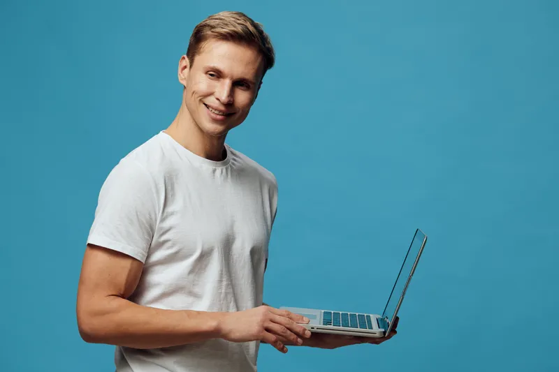 smiling man holding laptop wearing white t-shirt looking at camera digital nomad remote work technology blue background studio portrait
