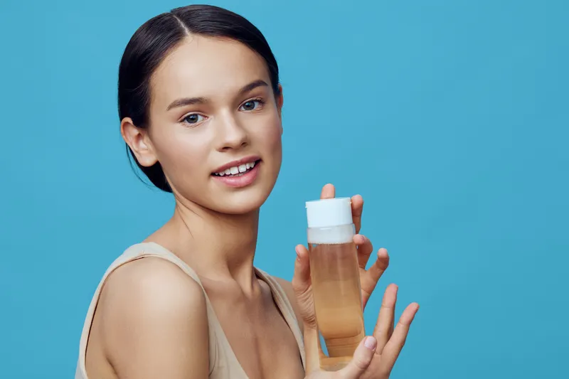 Young woman with long dark hair holding a skincare bottle, promoting healthy skin care on a vibrant blue background