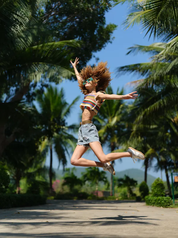 Energetic woman jumping outdoors in summer attire with vibrant colors and tropical background, expressing joy and freedom in bright daylight.