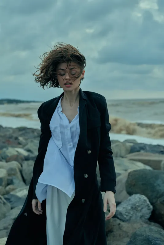 Windy beach scene with a woman in a long coat and light shirt, moody sky, rocky shore, atmospheric fashion moment by the sea