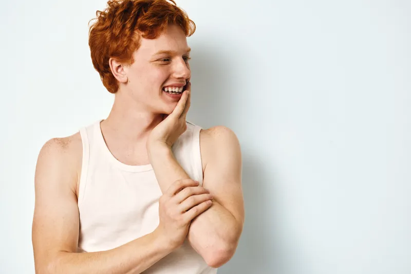 Young man with red hair and freckles wearing white sleeveless shirt smiling looking aside touching face and arm with happy expression on light background.
