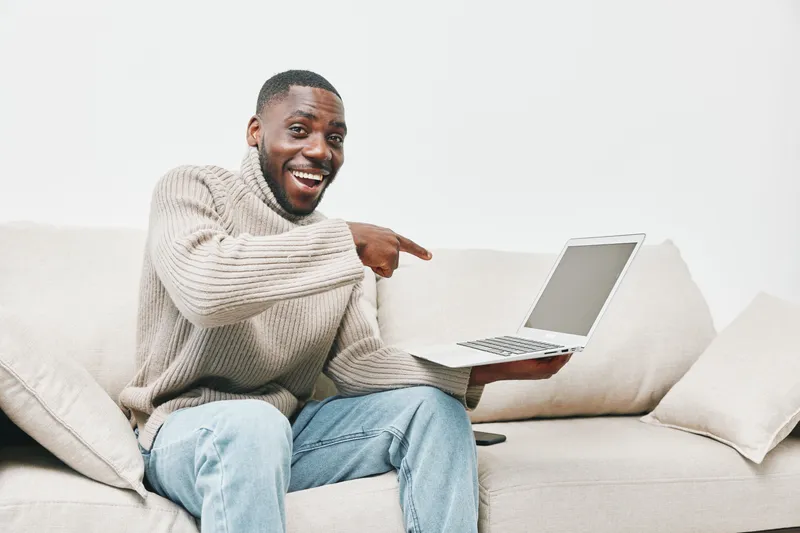 Happy young African man in cozy sweater showing laptop screen at home, with joyful expression and stylish casual outfit, conveying excitement about technology