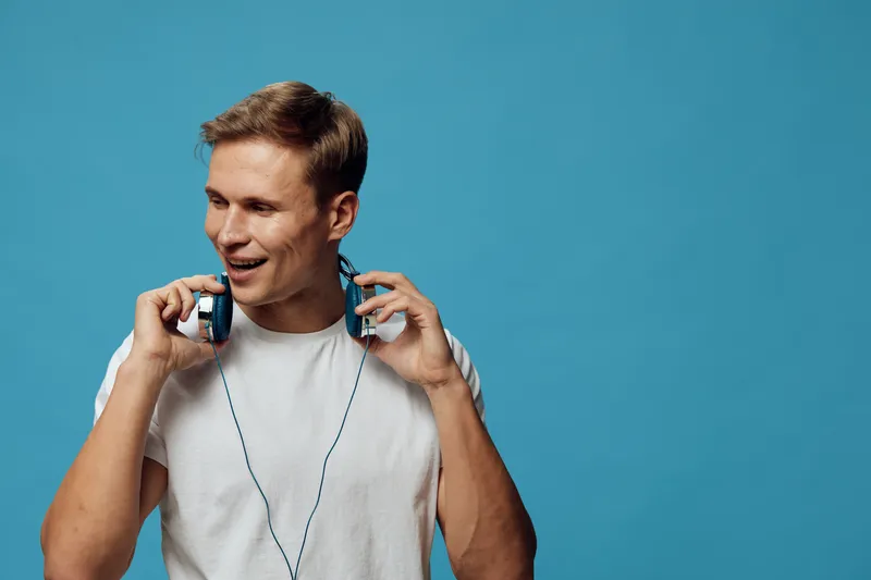 Smiling man with headphones, casual white T-shirt, holding earphones against a vibrant blue background, showcasing a joyful and relaxed mood. Perfect for lifestyle, music, or technology themes, vibran
