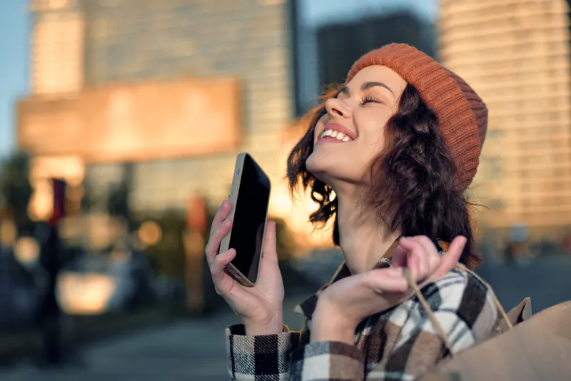 Woman city shopping phone smile portrait enjoying golden hour glow and candid lifestyle moment; authenticity and mindful living captured in emotional storytelling urban scene.