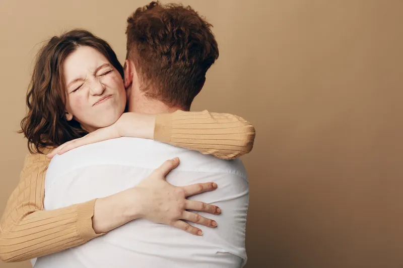 Young woman with closed eyes hugs man wearing white shirt, expressing affection and comfort. Intimate moment captured on beige background, concept of love and emotional connection.