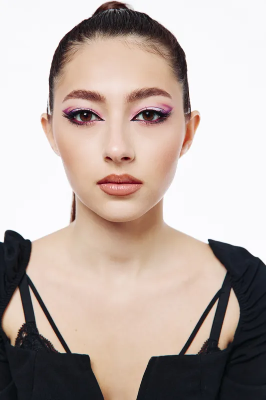 Young woman with creative pink eyeshadow and winged eyeliner stares confidently. She wears a stylish black top reflecting bold beauty and fashion concept. Studio light highlights her face.