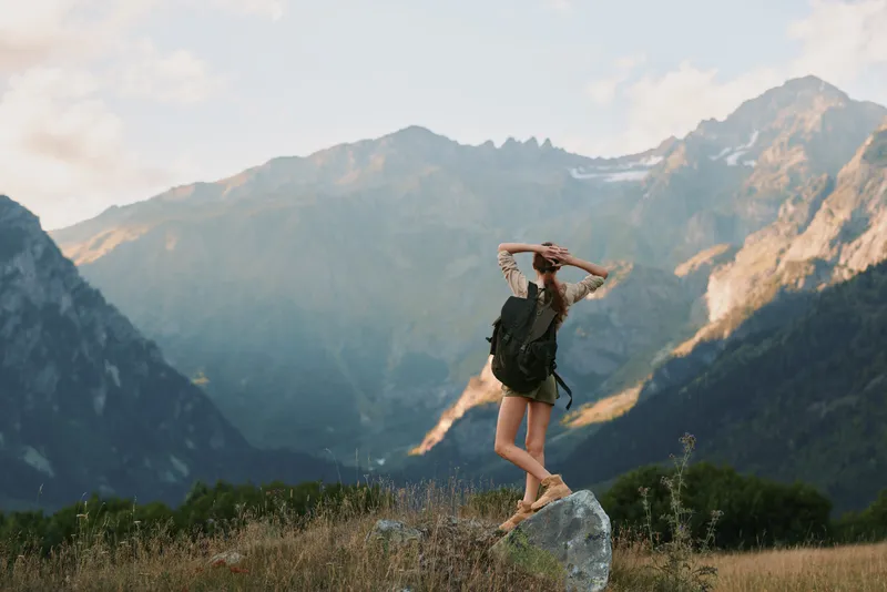 Young woman traveler with backpack enjoys mountain landscape standing on rock in nature, relaxing and stretching arms, lifestyle adventure concept during summer day.