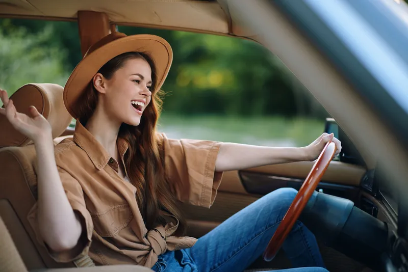 Joyful young woman driving a vintage car, wearing a stylish hat and a relaxed outfit, surrounded by vibrant greenery, evoking a sense of freedom and adventure.