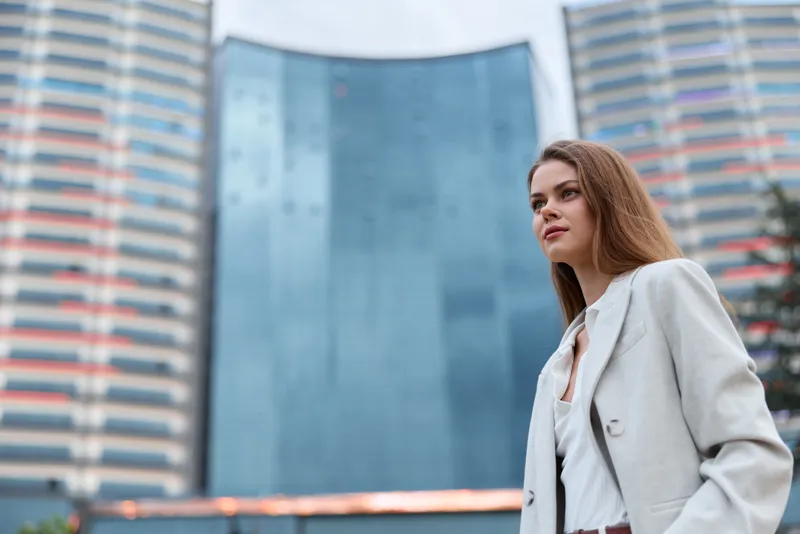 Professional business woman in light suit standing confidently in front of modern glass building with urban cityscape background