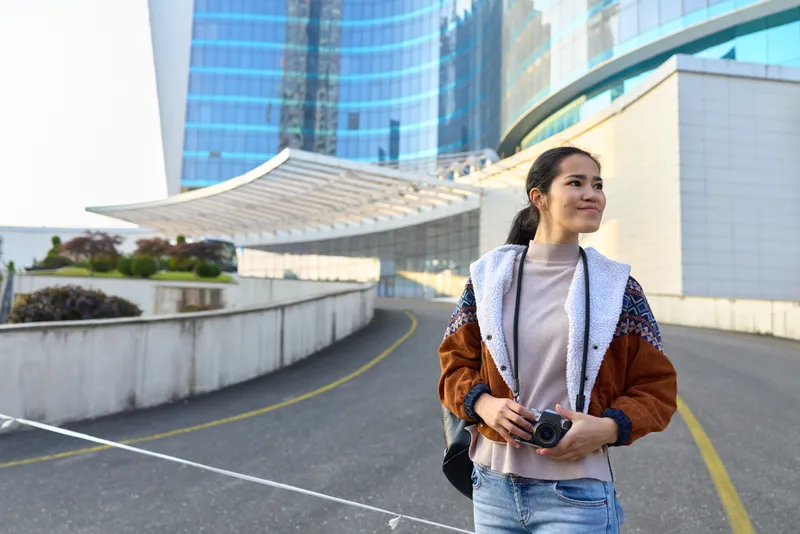 Woman holding a camera in a modern urban outdoor setting, stylish jacket, city skyline behind, ready for street photography and travel memories, relaxed pose, bold colors, lifestyle vibe