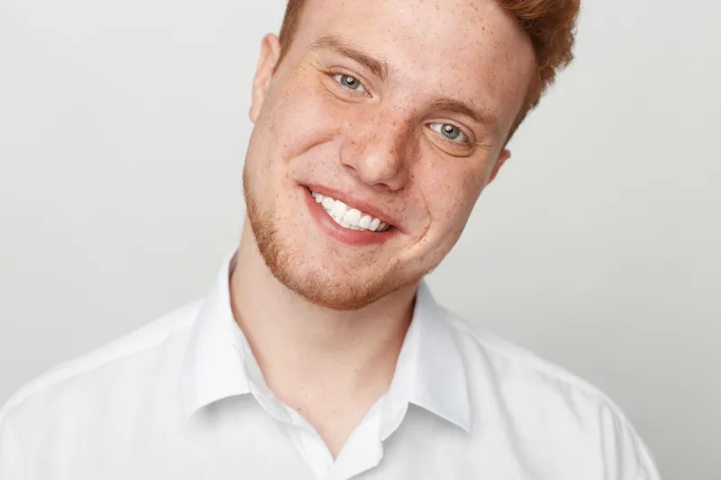 Smiling young man with red hair and freckles wears white shirt, looking at camera on light background. Concept of happiness, confidence, and natural beauty in portrait photography.