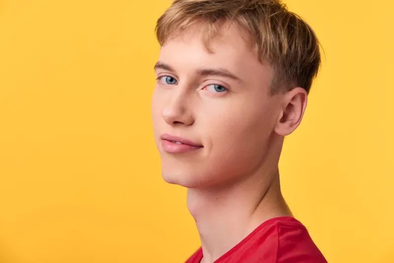 Young man with a subtle smile in a red shirt against a vibrant yellow background, close up shot highlighting clear skin and confident mood, suitable for lifestyle and fashion campaigns
