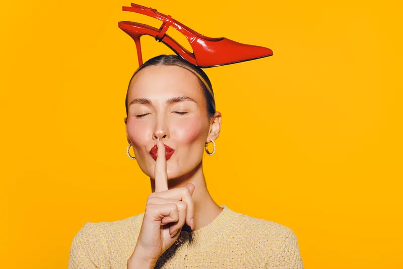 Woman with closed eyes holds finger to lips in shushing gesture, wearing red high heel on head against bright yellow background. Concept of silence, fashion, and playful expression.