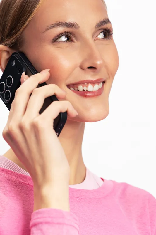 Smiling young woman in pink sweater holds smartphone to ear during call. Close-up on white background. Concept of communication, technology, and positive emotions in everyday life.