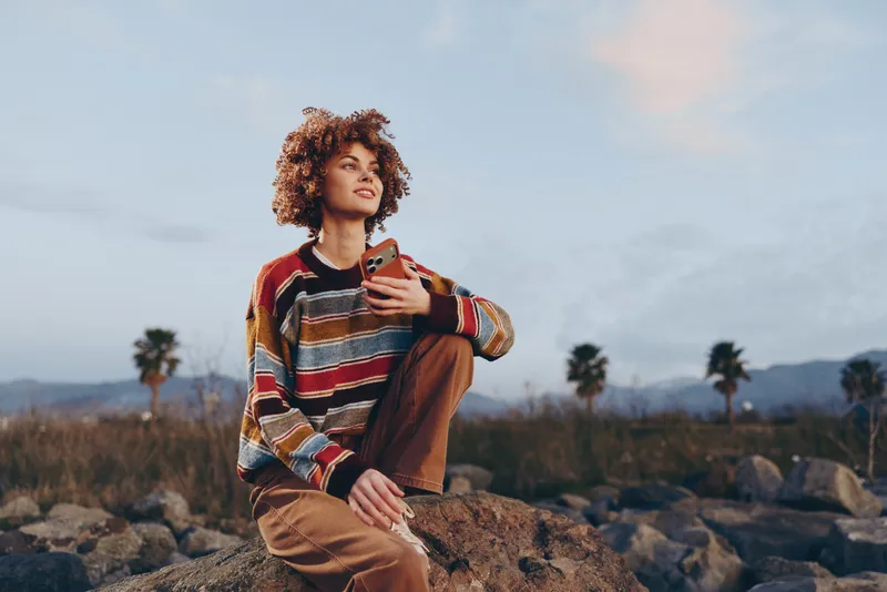 Woman lifestyle outdoors, curly haired woman sitting on rocks, casual striped sweater, warm sunset light, holding a small item, relaxed pose, palm trees and hills softly blurred