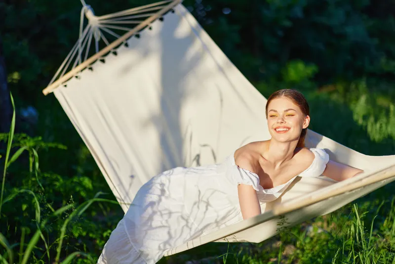 Young woman in a white dress relaxing in a hammock in a green garden, smiling joyfully, with soft sunlight highlighting her serene expression