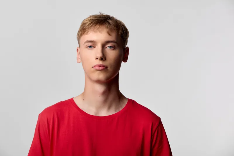 Young man wearing a red shirt stands in a studio with a neutral background, a serious expression and direct gaze, suitable for modern lifestyle and fashion campaigns