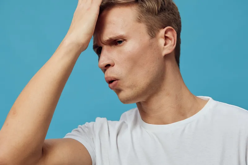Male young adult with short brown hair and light skin shows confusion or frustration holding head with hand, looking down, wearing casual white t-shirt, isolated on blue background, studio portrait, e