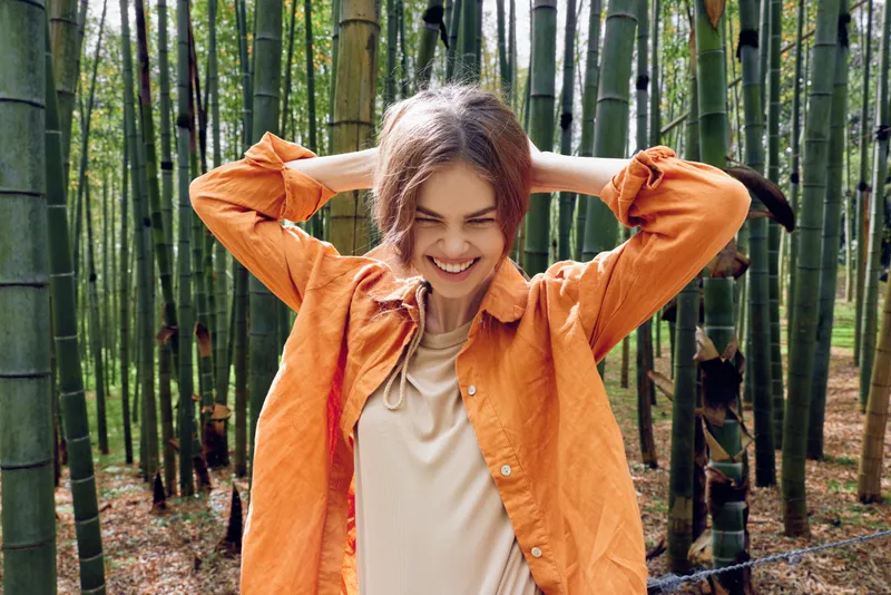 Woman bamboo forest smile orange portrait of a young woman in an orange jacket laughing with hands behind her head in a bamboo grove, candid nature travel portrait full of joy.