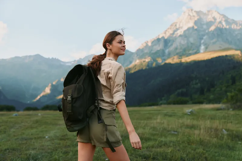 Young woman with backpack walks through a grassy meadow toward distant mountains, enjoying fresh air and open space, her relaxed expression conveying a carefree outdoor lifestyle.